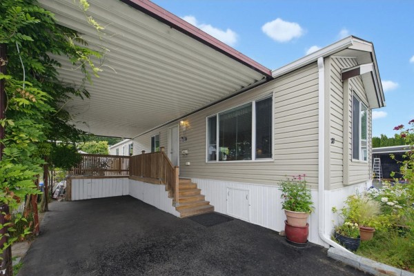 Front view of a beige mobile home with a carport, flower garden, and asphalt driveway. - 38 15875 20 Avenue, Surrey