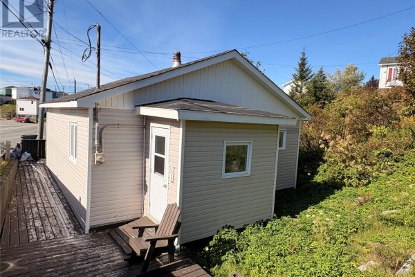 Rear exterior of a beige bungalow with a small deck, wooden chairs, and natural foliage. - 38 Brook Street, Port Aux Basques