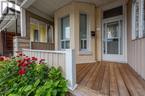 Close-up of the covered front porch entrance with wooden decking, beige siding, and red flowers. - 38 Gordon Street, Toronto