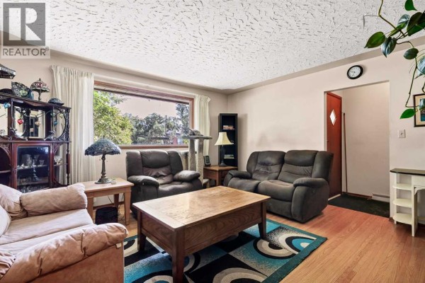 Living room with beige sofas, two recliner chairs, a wooden coffee table, and a large window overlooking greenery. - 3803 4 Avenue Sw, Calgary