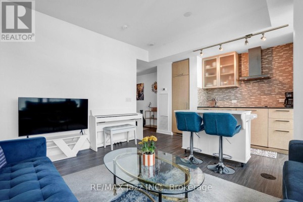 Living room area with white piano, TV stand, dark wood floors, and view of kitchen. - 3808 - 14 York Street, Toronto