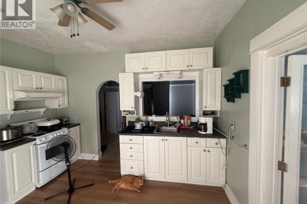 Kitchen showing white cabinets, stove, sink, window with blue curtains, and doorway. - 381 Main Street, Bengough