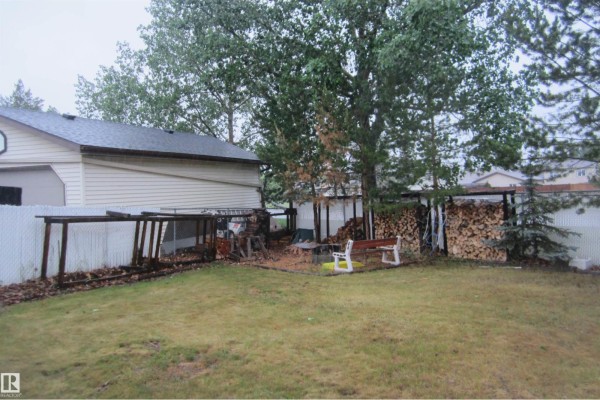 Backyard with a green lawn, white fence, wooden sheds, and stacked firewood. - 3814 37 Av, Leduc