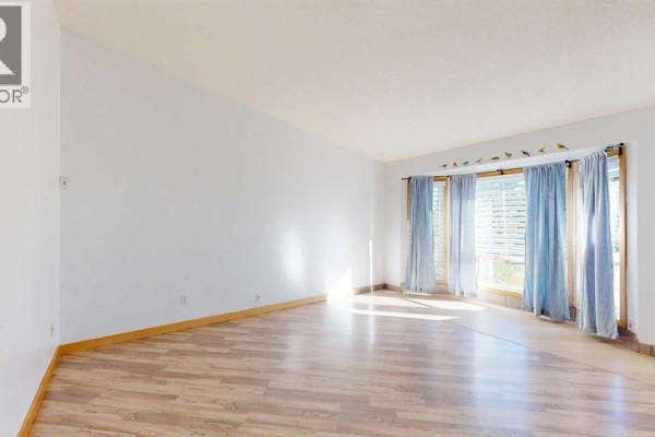 Empty living room with light wood laminate flooring and large windows with blue curtains. - 3818 61a Street, Stettler