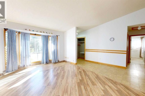 Living room with wood laminate flooring, large windows, blue curtains, and view of hallway closet. - 3818 61a Street, Stettler
