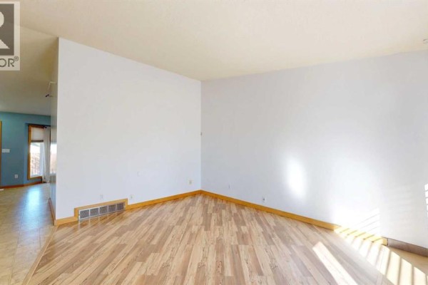 Empty corner of a living room with light wood laminate flooring and a floor vent. - 3818 61a Street, Stettler