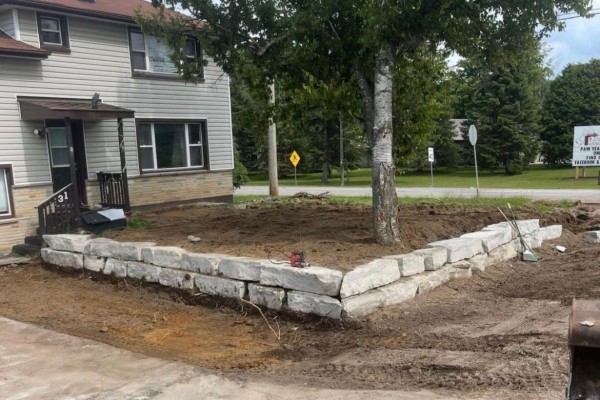 Stone retaining wall bordering a dirt landscaping area with a large tree in front of a beige building. - 3831 Soules Road, Severn
