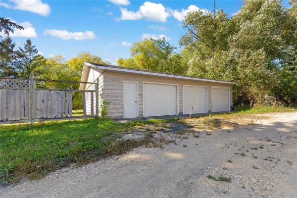 Three-bay detached garage with white doors, located on a gravel driveway with a wooden fence to the left. - 39 First Avenue N, Blumenort