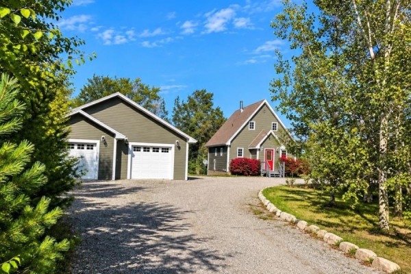 A gravel driveway leads past a two-car garage and dark green house surrounded by trees. - 39 Main Road, Town Of Terra Nova