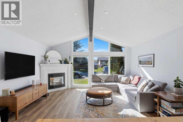 Living room with a sectional sofa facing large corner windows, a fireplace, and a television mounted on the wall. - 39 Midridge Close Se, Calgary