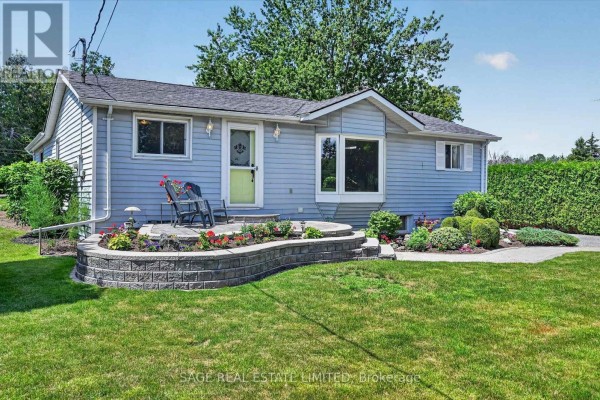 Side view of a light blue bungalow with a paved driveway and mature trees in the yard. - 39 Riverside Drive, Kawartha Lakes