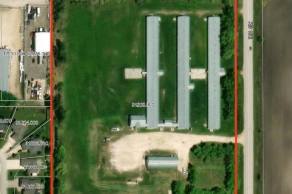 Aerial view of 390 Main Street, Niverville showing a large property with multiple long, grey buildings and an open field. - 390 Main Street, Niverville