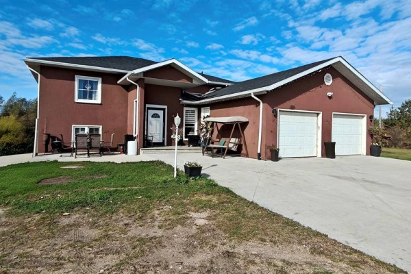 Exterior of a large brown stucco house with two white garage doors and a light-colored driveway. - 39044 Road 34n Road, La Broquerie