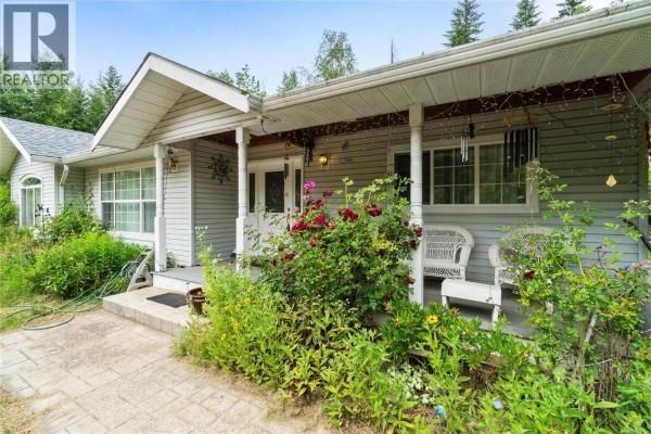 Close-up of the covered front porch with white wicker chairs and flowering bushes in the foreground. - 3915 Express Point Road, Scotch Creek