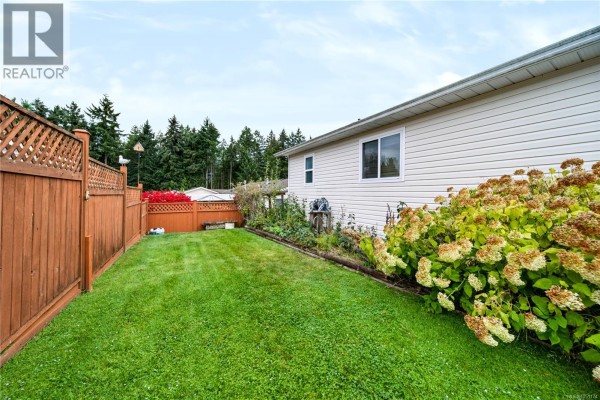 Backyard of a beige mobile home with a green lawn and a wooden privacy fence. - 3931 Excalibur St, Nanaimo