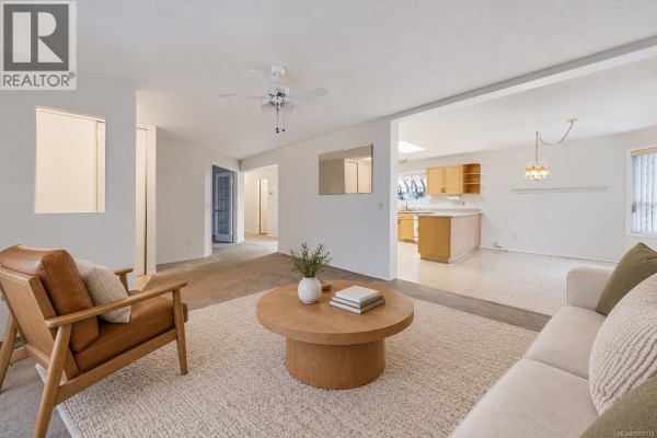 Living room with tan armchair, beige sofa, round wooden coffee table, and view of kitchen. - 3931 Excalibur St, Nanaimo