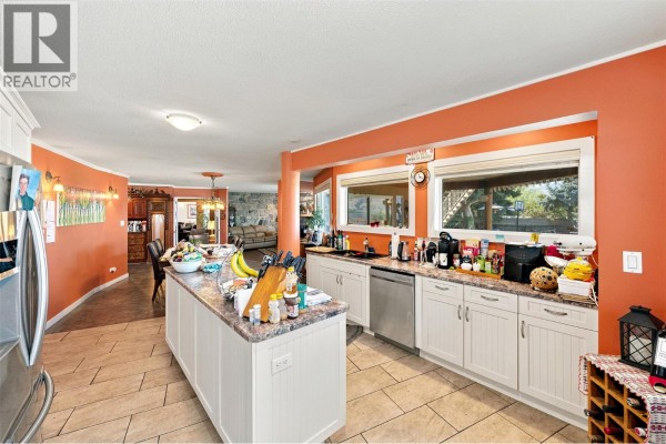 Wide shot of the kitchen with an island, white cabinets, and views through windows. - 3935 Lakeside Road, Penticton