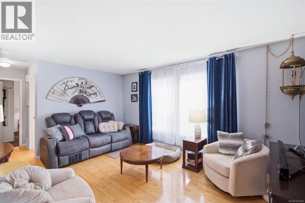 Living room with grey reclining sofa, wood coffee table, armchair, and blue curtains. - 3948 Valewood Dr, Nanaimo