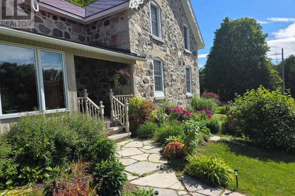 Close-up of a stone farmhouse entrance with a covered porch and colorful flower gardens. - 395 Elsinore Road N, South Bruce Peninsula