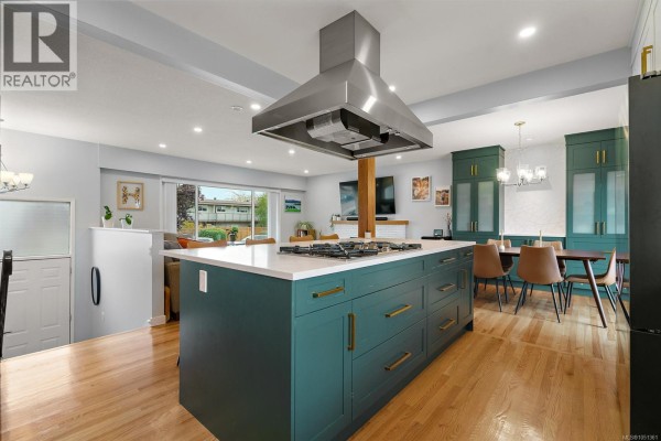 Open-concept kitchen and dining area with a teal island, wooden floors, and a view of the backyard. - 3988 Larchwood Dr, Saanich