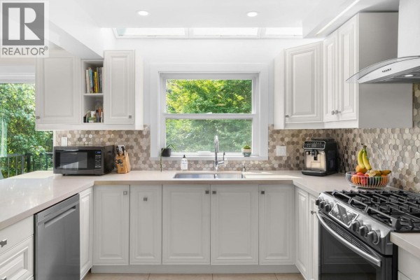 U-shaped kitchen with white shaker cabinets, a mosaic tile backsplash, stainless steel appliances, and a window overlooking trees. - 4 230 W 16th Street, North Vancouver