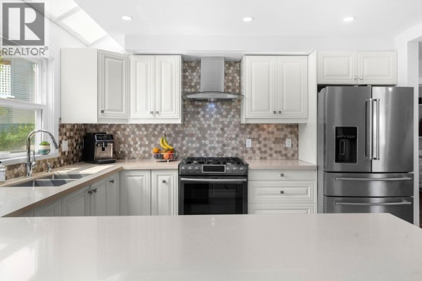 A kitchen island with a quartz countertop faces a stove, stainless steel refrigerator, and white cabinetry with mosaic tile backsplash. - 4 230 W 16th Street, North Vancouver