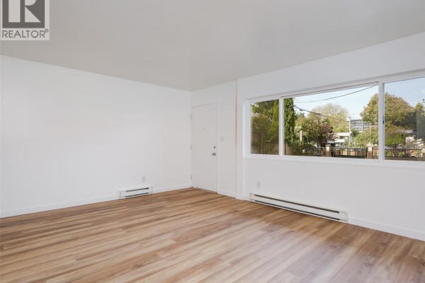 Living room with light wood laminate flooring, white walls, a textured accent wall, and a doorway to a kitchen. - 4 740 Wilson St, Victoria