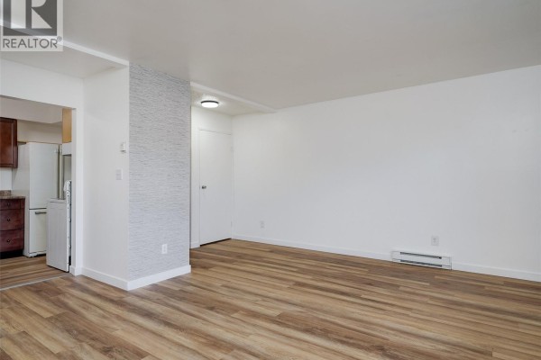Living room with light wood laminate flooring, white walls, an accent wall, and a view into a kitchen area. - 4 740 Wilson St, Victoria