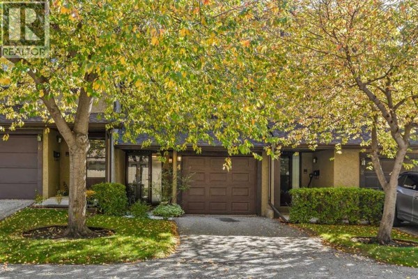 Exterior view of a townhouse with a brown garage door and trees in front. - 4 Glamis Gardens Sw, Calgary