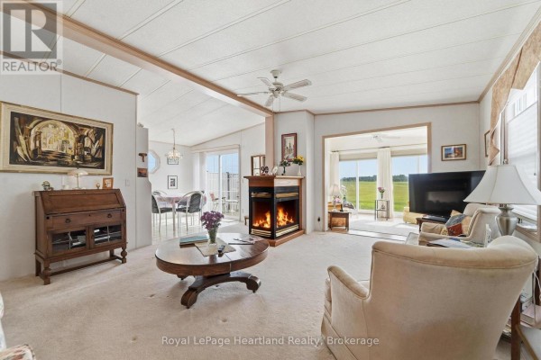 Living room with a fireplace, plush carpet, sofa, armchair, and a view of a dining area and field. - 40 Algonquin Lane, Ashfield Colborne Wawanosh