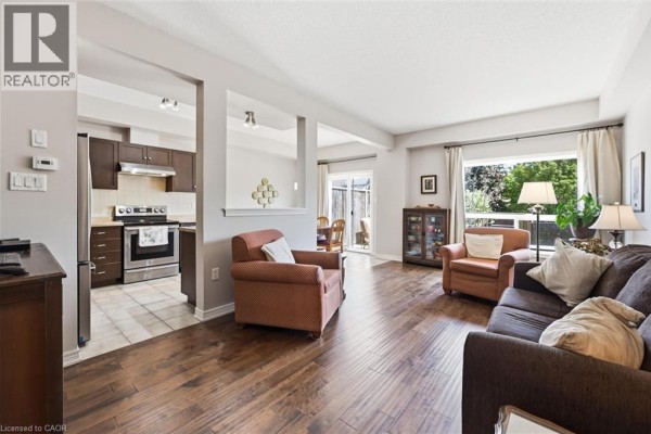 Living room with dark wood flooring, featuring a sofa, armchairs, and a partial view of the kitchen. - 40 Dartmouth Gate Unit# 10, Stoney Creek
