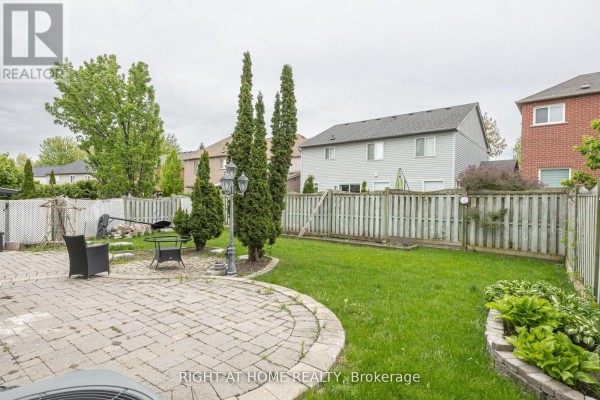 Backyard view with a circular stone patio, green grass, wooden fence, and trees. - Basement - 40 Sabbe Crescent, Ajax