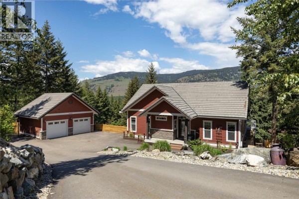 Red house with a two-car garage and a driveway surrounded by trees and mountains. - 4006 Coachwood Close, Coldstream