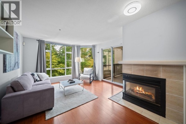 Living room with grey sofa, armchair, coffee table, fireplace, and large bay window. - 401 22233 River Road, Maple Ridge