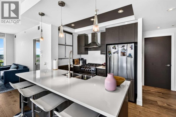 Kitchen with a large white island, stainless steel appliances, dark cabinets, and a view of the living area. - 402, 10 Shawnee Hill Sw, Calgary
