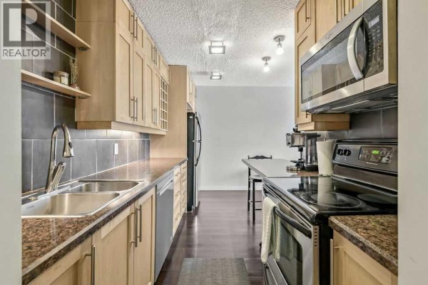 Galley kitchen with light wood cabinets, stainless steel sink, and dark tile backsplash. - 402, 2140 17a Street Sw, Calgary
