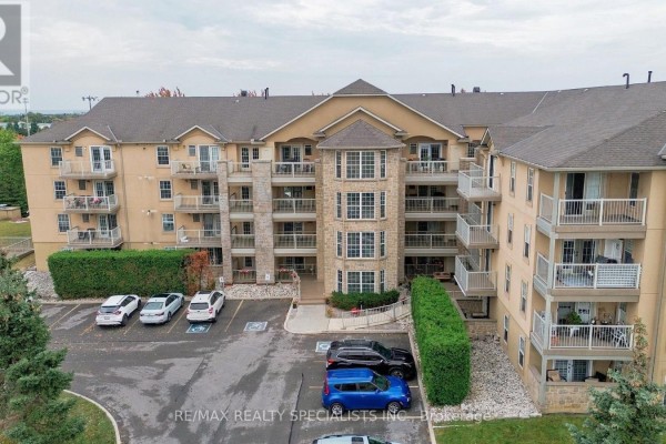 Exterior view of a beige apartment building with balconies, entrance, and surrounding parking lot. - 402 - 1470 Bishops Gate, Oakville