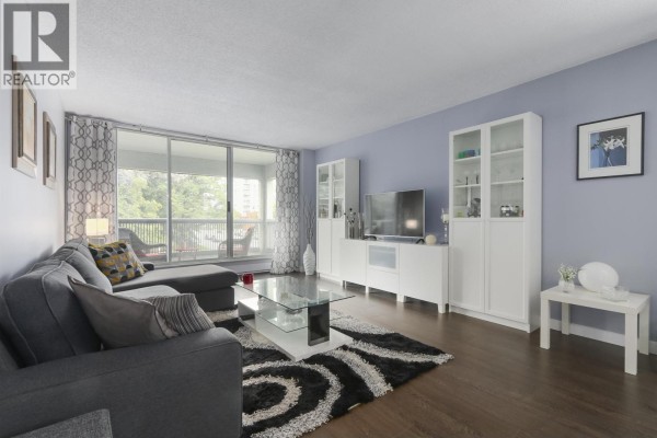 Living room with a grey sectional sofa, glass coffee table, and white media units. - 402 550 Eighth Street, New Westminster