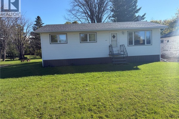 Front view of a white vinyl-sided bungalow with a lawn, trees, and a picnic table. - 403 & 401 Main Street, Pelly