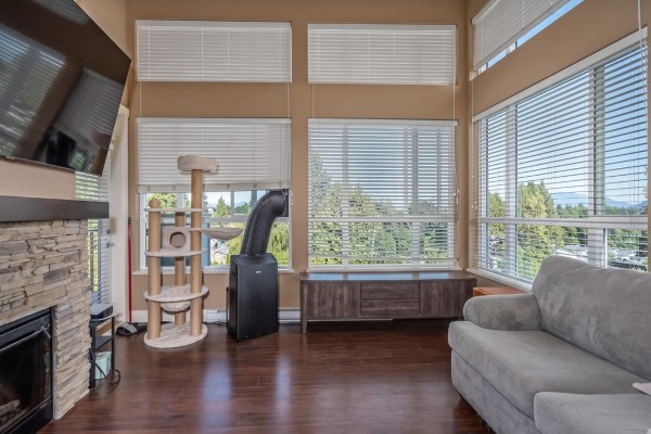 Living room with fireplace, large windows, sofa, and view of trees and mountains. - 403 3192 Gladwin Road, Abbotsford