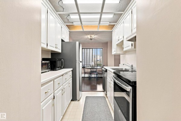 Long galley kitchen with white cabinets, black appliances, and a view of a dining nook. - #403 9725 106 St Nw, Edmonton