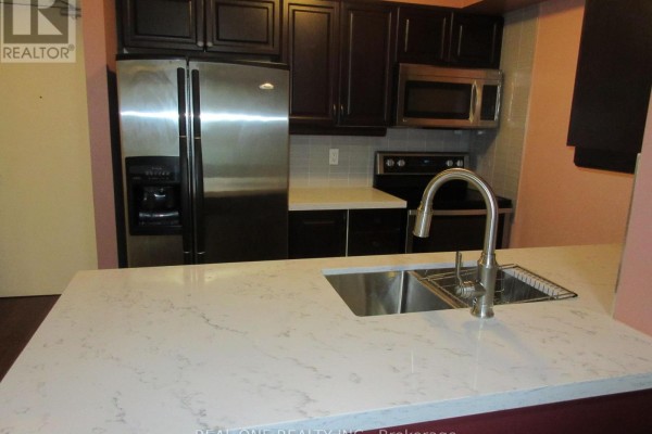 Close-up of kitchen counter with double sink and faucet, stainless steel refrigerator, and dark wood cabinets. - 404 - 61 Town Centre Court, Toronto