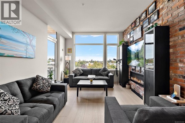 Living room with grey sectional sofa, coffee table, large windows, and exposed brick accent wall. - 404 1216 Carlisle Ave, Esquimalt