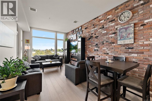 Living and dining area with grey sofas, dark wood table, chairs, and exposed brick wall. - 404 1216 Carlisle Ave, Esquimalt