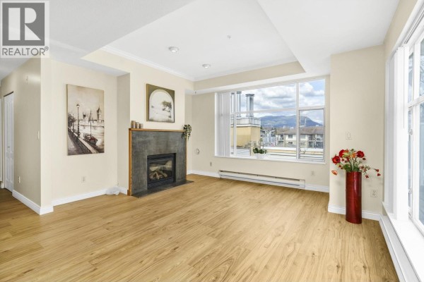 Living room showing fireplace, doorway to kitchen, and beige walls with light wood floors. - 404 2340 Hawthorne Avenue, Port Coquitlam