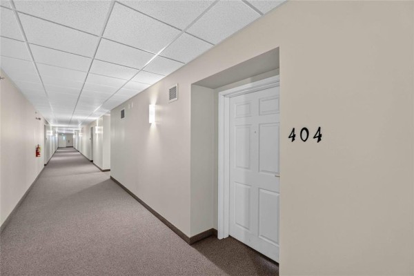 Long, carpeted interior hallway of an apartment building with multiple doors and a drop ceiling. - 404 700 Allegheny Drive, Winnipeg