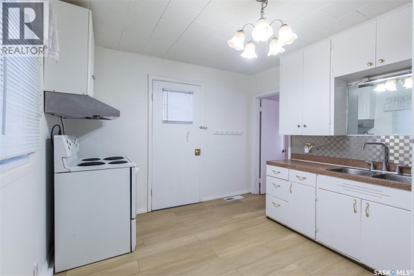 A white kitchen with a white stove, white cabinets, double sink and a door. - 404 Fairford Street W, Moose Jaw