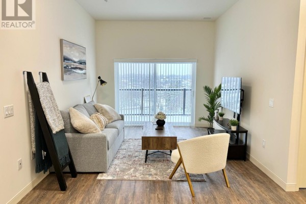 Living room with grey sofa, coffee table, armchair, television, and sliding glass door with vertical blinds. - 405 - 135 Station Street, Belleville