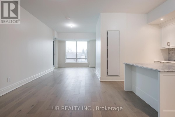 Living area with large window overlooking trees, light wood floors, and a view of the kitchen. - 405 - 1451 Wellington Street W, Ottawa