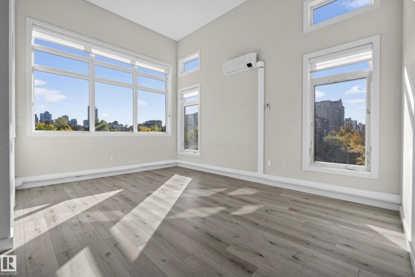 Empty room with light wood floors and multiple windows offering city views. - #405 10227 115 St Nw, Edmonton